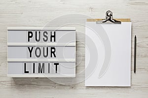 `Push your limit` words on a lightbox, clipboard with blank sheet of paper on a white wooden surface, top view. Overhead, from