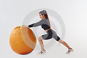 Pushing giant orange. Sportive woman in black clothes in the studio against white background