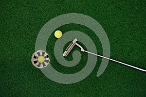 Putting golf club on green grass with golf ball in the hole - top view.