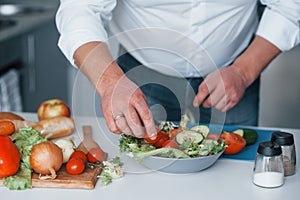 Putting little tomatoes in salad. Man in white shirt preparing food on the kitchen using vegetables