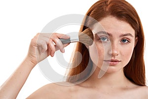 Putting some makeup. Portrait of half naked cute red-haired woman with freckles holding makeup brush and looking at