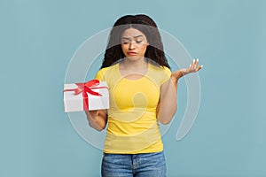 Puzzled african american woman holding gift decorated with ribbon, standing over blue background, studio shot