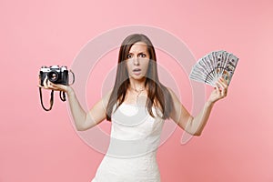 Puzzled bride woman in wedding dress holding retro vintage photo camera, bundle lots of dollars, cash money choosing