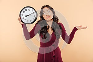 Puzzled brunette woman with curly long hair holding clock showing time after 8 gesturing like she is late or do not care over pea