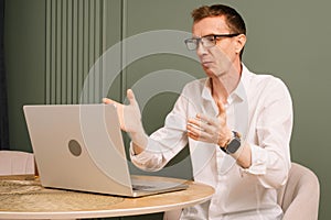 Puzzled business man sit at table, looking at laptop screen with misunderstood eyes. Dazed human raised hands in shock.