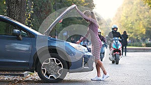 Puzzled female driver standing on a street near her car with popped up hood inspecting broken engine.