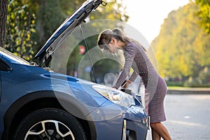 Puzzled female driver standing on a street near her car with popped up hood inspecting broken engine