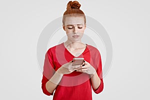 Puzzled freckled young woman with natural red hair tied in knot, wears casual shirt, looks in cell phone with displeased expressio