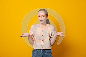 Puzzled girl, spreads hands with doubt,over yellow background. Teen girl faces dilemma, being unsure