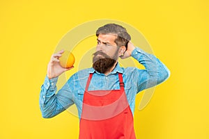 Puzzled man in apron looking at fresh orange fruit scratching head yellow background, fruiterer photo