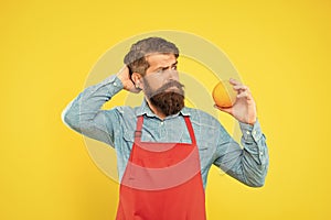 Puzzled man in apron looking at fresh orange fruit scratching head yellow background, fruiterer