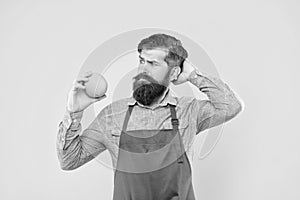 Puzzled man in apron looking at fresh orange fruit scratching head yellow background, fruiterer photo