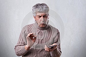 A puzzled mature man in brown sweater holding smartphone trying to understand how to switch it on. Troubled elderly man holding sm