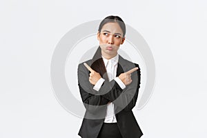 Puzzled pretty asian businesswoman in black suit troubled making choice, standing indecisive, pointing fingers sideways