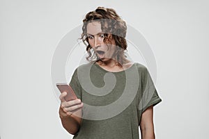 Puzzled stupefied curly hair woman opens eyes and mouth widely holding smartphone
