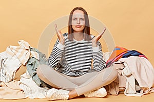 Puzzled unhappy Caucasian woman posing near heap of multicolored unsorted clothes isolated over beige background sitting with