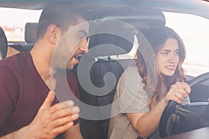 Puzzled young female driver trainee drives car, listens attentively male instructor who gives some advice, shows how to do it righ