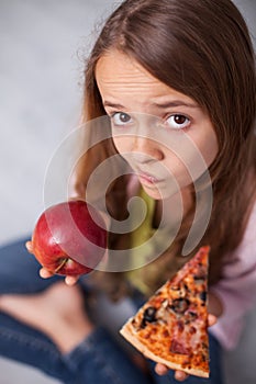 Puzzled young girl undecided wheter to choose the appetizing pizza or healthy apple