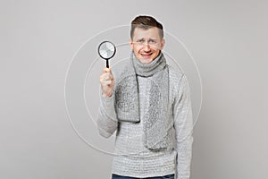 Puzzled young man in gray sweater, scarf holding magnifying glass on grey wall background in studio. Healthy