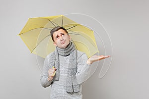 Puzzled young man in gray sweater, scarf holding yellow umbrella, pointing hand aside, looking up on grey