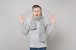 Puzzled young man in gray sweater, scarf spreading hands showing palms isolated on grey background in studio. Healthy