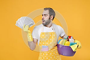Puzzled young man househusband in apron rubber gloves hold basin with detergent bottles washing cleansers doing
