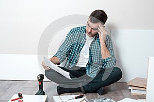 Puzzled young man sitting on the floor, reading the instructions for the Assembly of furniture