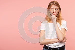 Puzzled young pretty long haired redhead lady holding her chin with raised hand and looking pensively aside, wearing casual