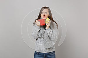 Puzzled young woman in gray sweater, scarf holding lemon, red cup of tea isolated on grey wall background. Healthy