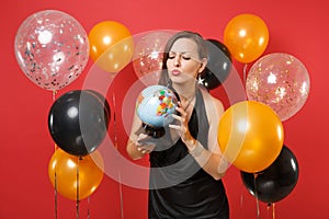 Puzzled young woman in little black dress celebrating, looking on world globe in hands on red background air balloons