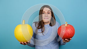 Puzzled young woman shrugs shoulders, has uncertain facial expression, can`t choose which pumpkins, wears sweater