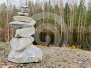 A pyramid of stones stands on a stone against the background of a river, beyond which a coniferous forest can be seen.