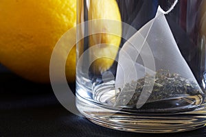 A pyramid tea bag with green tea in a transparent mug next to a fresh yellow lemon. Macro. Black style