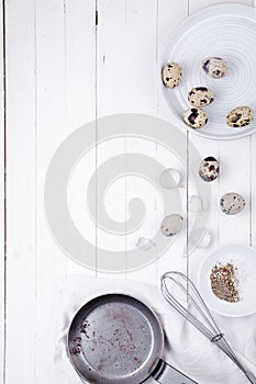 Quail eggs in a plate, a shell, a whisk for beating and a frying pan on a white wooden background. view from above