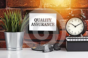 QUALITY ASSURANCE text with alarm clock, books and vase on brick background