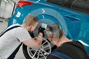 Quality with new standards. Two male mechanics using torch for inspecting wheel of lifted car at auto repair shop