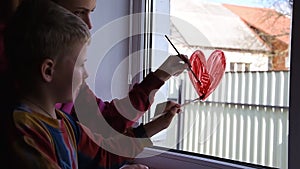 Quarantine. Mother with boy draws red heart on the window with paint. Waiting for the rainbow, Stay home