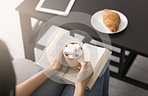 Quarantine pastimes. Above view of young girl with open book and cup of coffee with marshmallows at home