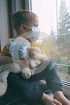 Quarantine, threat of coronavirus. Sad child and his teddy bear both in protective medical masks sits on windowsill and looks out