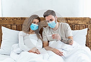 Quarantine together. Sad young man and woman in protective masks look at thermometer