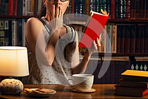 Quarantine. A young Caucasian woman with glasses reads a book with fright. The home library is in the background. Close up.The