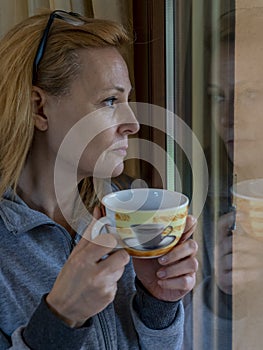 Quarantined blonde woman drinks a cup of tea looking at the outside world from the window in which it is reflected