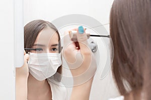 Quarantined life. A woman in a medical mask does makeup, mascaras her eyes. Prevention and protection against diseases