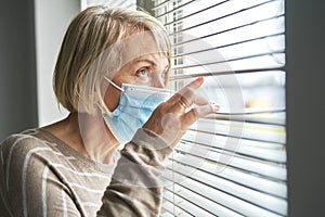 Quarantined senior woman wearing mask looks through blinds photo