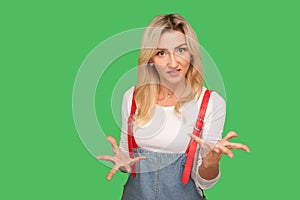 Quarrel, aggressive emotions. Portrait of irritated adult blond woman in stylish denim overalls raising her hands in anger