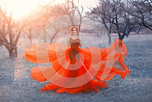 Queen in bright long elegant flying red dress poses for photo, woman with dark hair and crown on her head puts hands on