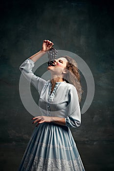 Queen. Vertical portrait of glamor young woman wearing blue historical dress going to take abite of grape. Food mood