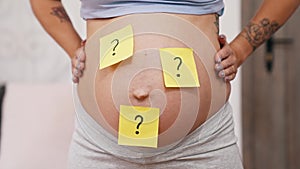 Question mark sticky notes on white pregnant woman's belly. Hands on hips. Unknown baby gender. Surprise. Horizontal