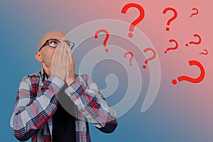 Question marks around a young man with glasses covering his face with his hands, psychological concept