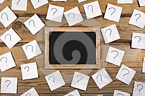 Question marks on crumpled note paper around a small blank blackboard on a wooden background